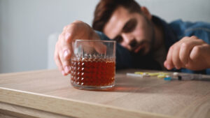 Uomo con capo reclinato, bicchiere di alcool in mano, siringhe e capsule su un tavolo, simbolo della dipendenza da alcool e droghe.